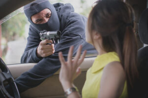 A woman sits in her car at an isolated road intersection, alarmed as an armed assailant points a gun at her while gripping the steering wheel, highlighting the tension and danger of a carjacking incident in a secluded area.
